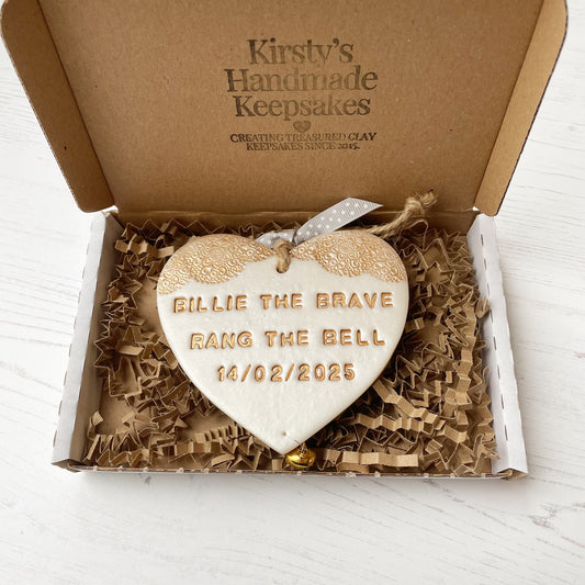 Personalised cancer survivor gift, pearlised white clay hanging heart with a gold lace edge at the top of the heart and a gold bell hanging below, the heart is personalised with BILLIE THE BRAVE RANG THE BELL 14/02/2025