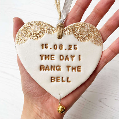 Personalised cancer survivor gift, pearlised white clay hanging heart with a gold lace edge at the top of the heart and a gold bell hanging below, the heart is personalised with 15.08.25 THE DAY I RANG THE BELL