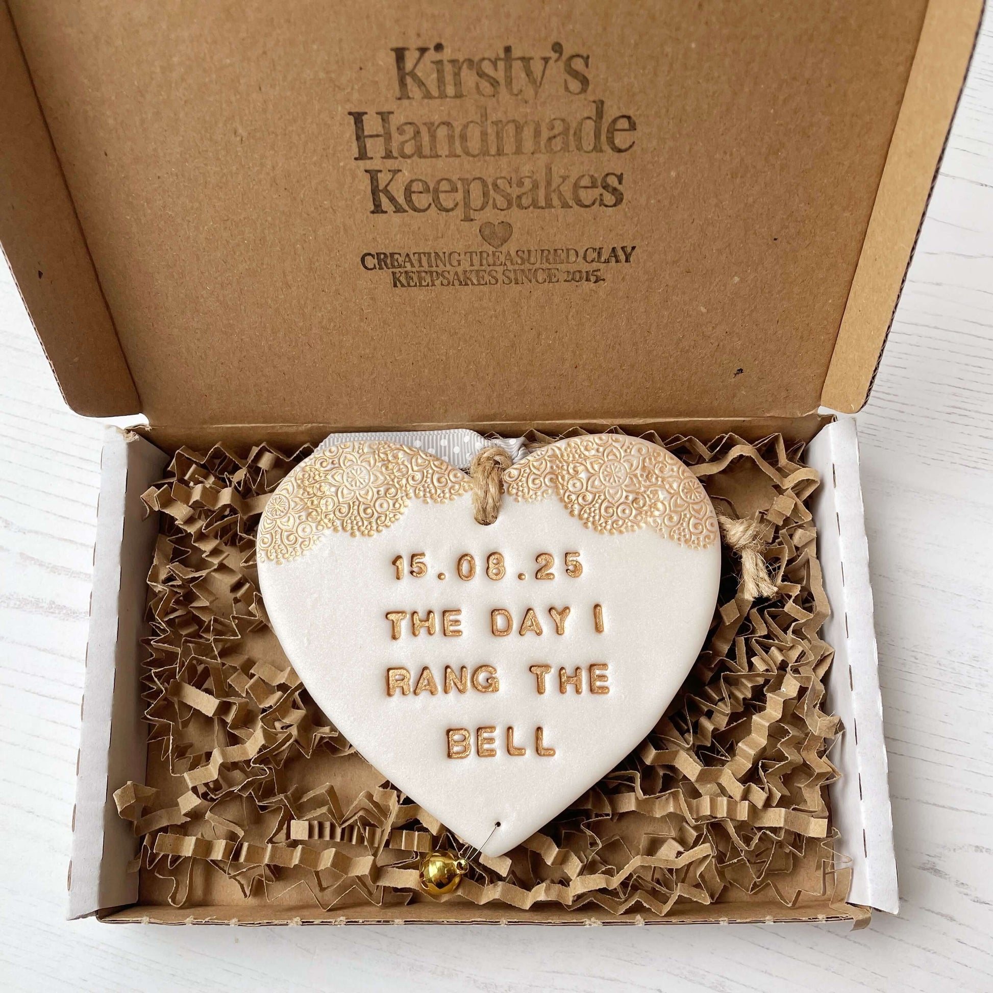 Personalised cancer survivor gift, pearlised white clay hanging heart with a gold lace edge at the top of the heart and a gold bell hanging below, the heart is personalised with 15.08.25 THE DAY I RANG THE BELL