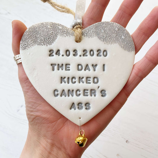 Personalised cancer survivor gift, pearlised white clay hanging heart with a grey lace edge at the top of the heart and a gold bell hanging below, the heart is personalised with 24.03.2020 THE DAY I KICKED CANCER’S ASS