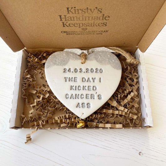 Personalised cancer survivor gift, pearlised white clay hanging heart with a grey lace edge at the top of the heart and a gold bell hanging below, the heart is personalised with 24.03.2020 THE DAY I KICKED CANCER’S ASS