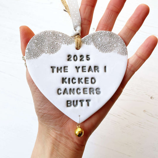 Personalised cancer survivor gift, glitter white clay hanging heart with a grey lace edge at the top of the heart and a gold bell hanging below, the heart is personalised with 2025 THE YEAR I KICKED CANCERS BUTT