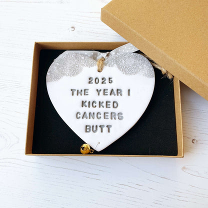 Personalised cancer survivor gift, glitter white clay hanging heart with a grey lace edge at the top of the heart and a gold bell hanging below, the heart is personalised with 2025 THE YEAR I KICKED CANCERS BUTT