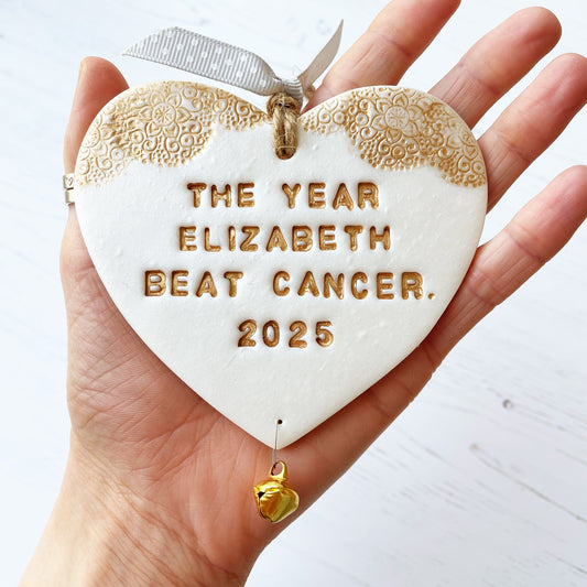 Personalised cancer survivor gift, pearlised white clay hanging heart with a gold lace edge at the top of the heart and a gold bell hanging below, the heart is personalised with THE YEAR ELIZABETH BEAT CANCER. 2025