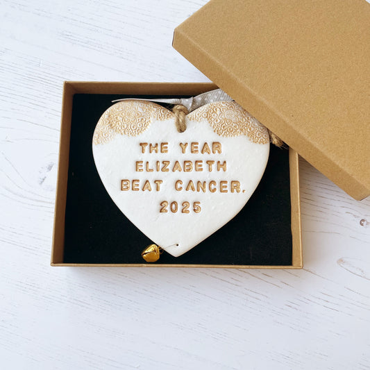 Personalised cancer survivor gift, pearlised white clay hanging heart with a gold lace edge at the top of the heart and a gold bell hanging below, the heart is personalised with THE YEAR ELIZABETH BEAT CANCER. 2025