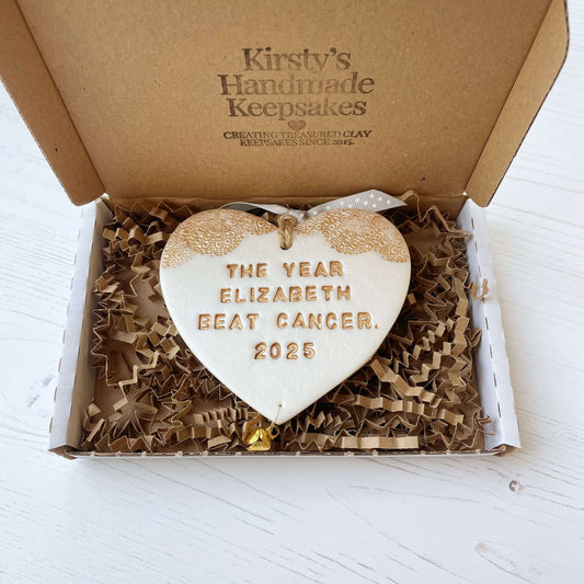 Personalised cancer survivor gift, pearlised white clay hanging heart with a gold lace edge at the top of the heart and a gold bell hanging below, the heart is personalised with THE YEAR ELIZABETH BEAT CANCER. 2025