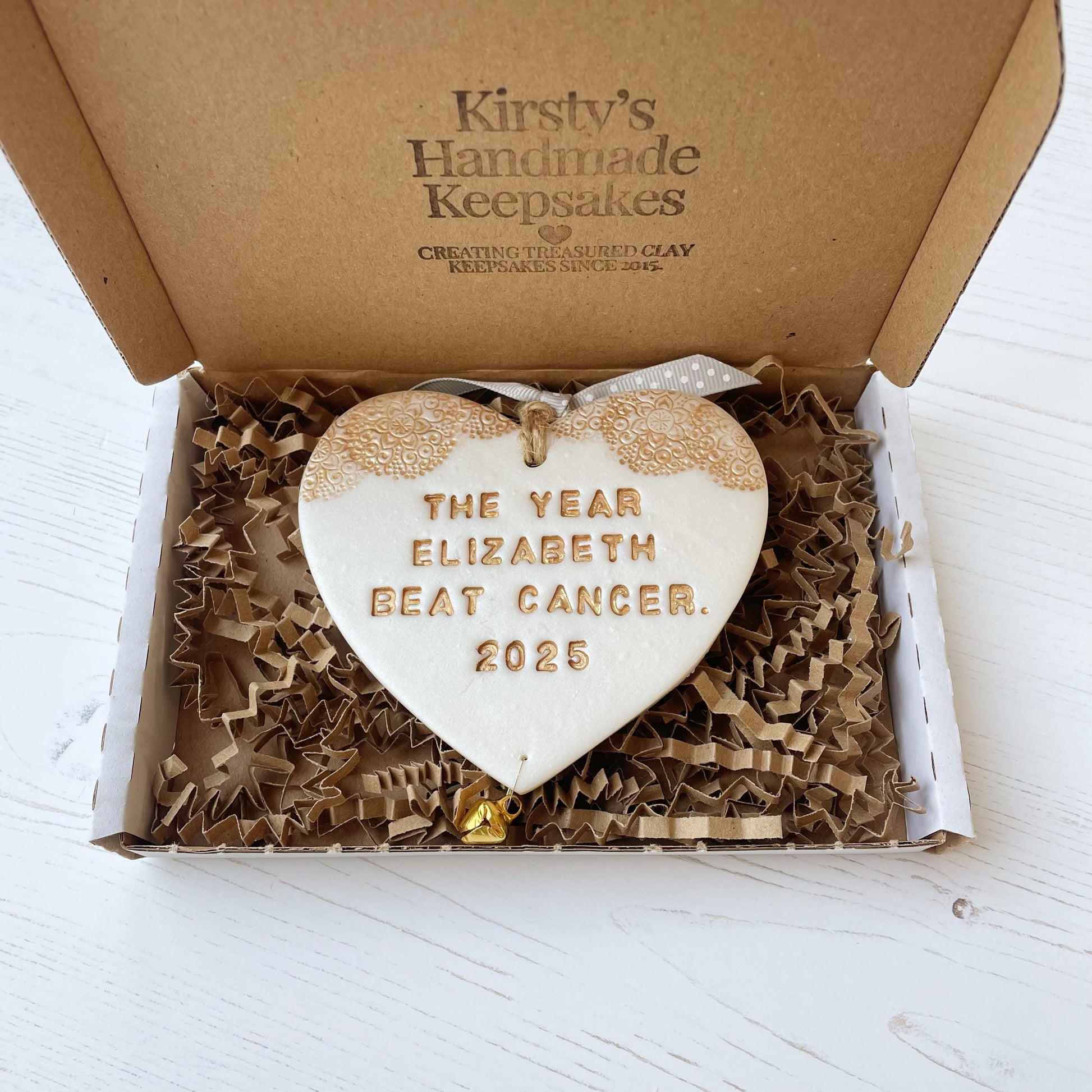 Personalised cancer survivor gift, pearlised white clay hanging heart with a gold lace edge at the top of the heart and a gold bell hanging below, the heart is personalised with THE YEAR ELIZABETH BEAT CANCER. 2025