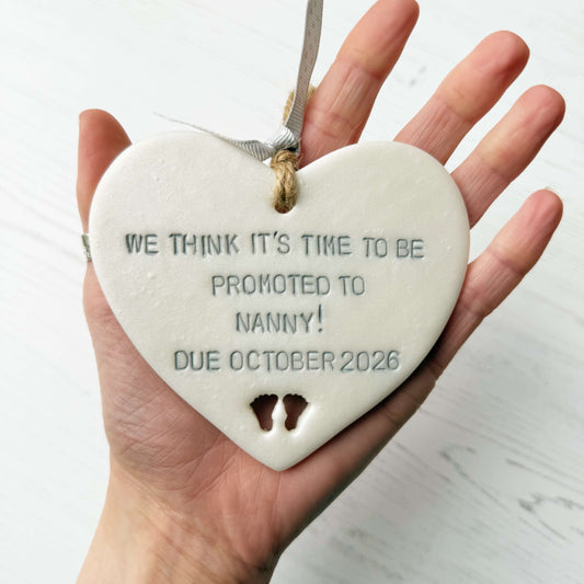 Pearlised white clay hanging heart with baby feet cut out of the bottom and grey personalisation, the heart is personalised with WE THINK IT’S TIME TO BE PROMOTED TO NANNY! DUE OCTOBER 2026