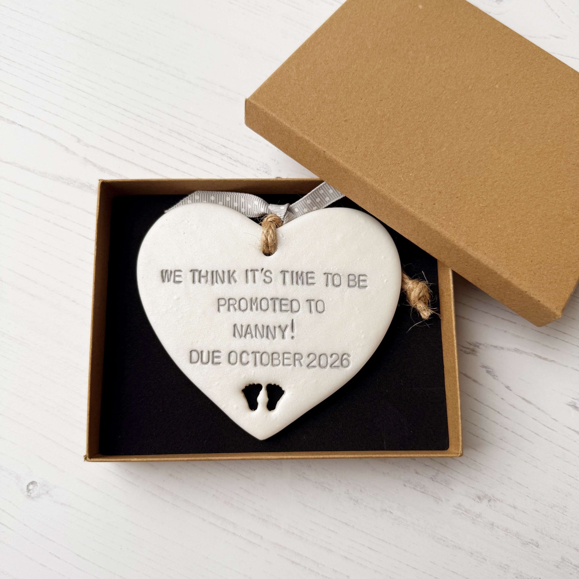 Pearlised white clay hanging heart with baby feet cut out of the bottom and grey personalisation, the heart is personalised with WE THINK IT’S TIME TO BE PROMOTED TO NANNY! DUE OCTOBER 2026