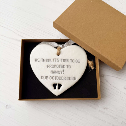 Pearlised white clay hanging heart with baby feet cut out of the bottom and grey personalisation, the heart is personalised with WE THINK IT’S TIME TO BE PROMOTED TO NANNY! DUE OCTOBER 2026
