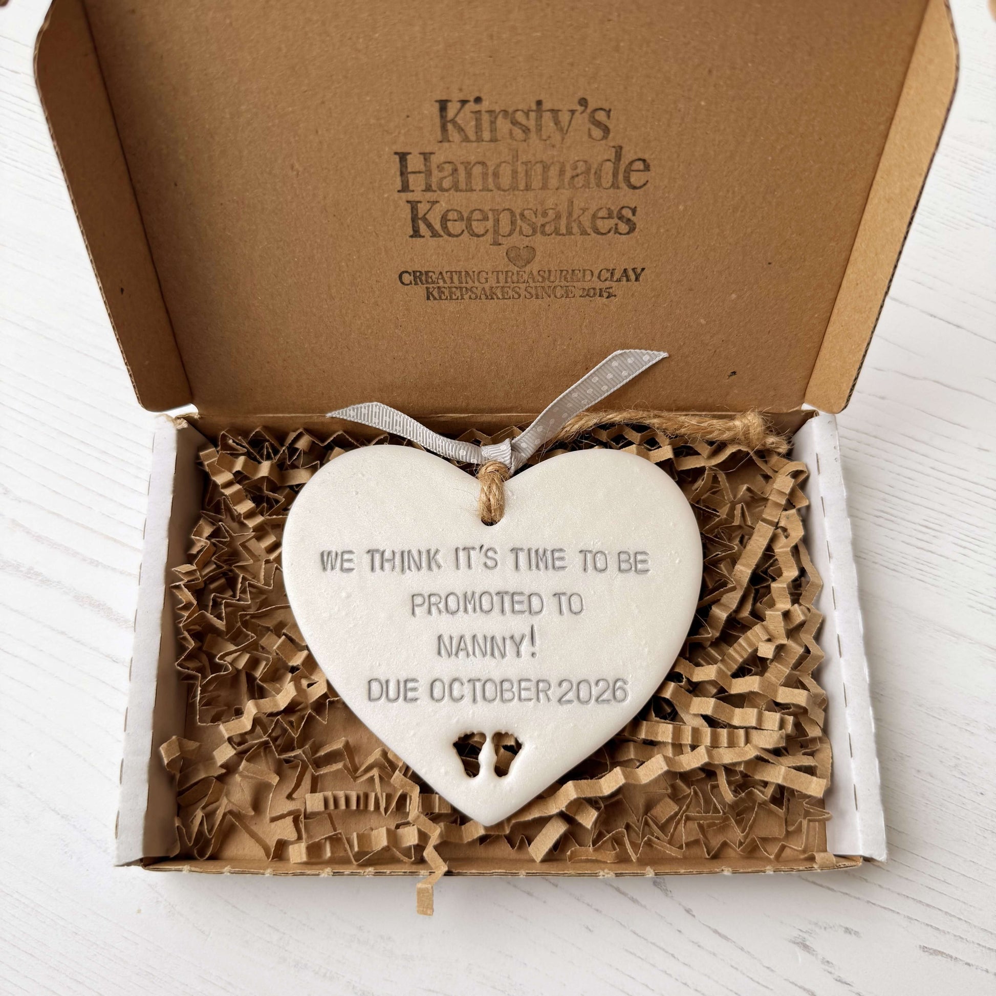Pearlised white clay hanging heart with baby feet cut out of the bottom and grey personalisation, the heart is personalised with WE THINK IT’S TIME TO BE PROMOTED TO NANNY! DUE OCTOBER 2026