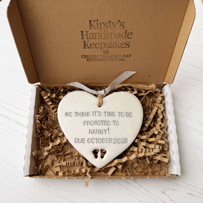 Pearlised white clay hanging heart with baby feet cut out of the bottom and grey personalisation, the heart is personalised with WE THINK IT’S TIME TO BE PROMOTED TO NANNY! DUE OCTOBER 2026