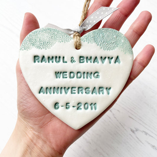 Personalised 13th wedding anniversary gift, pearlised white clay hanging heart with a sage green lace edge at the top of the heart, the heart is personalised with RAHUL & BHAVYA WEDDING ANNIVERSARY 6-5-2011