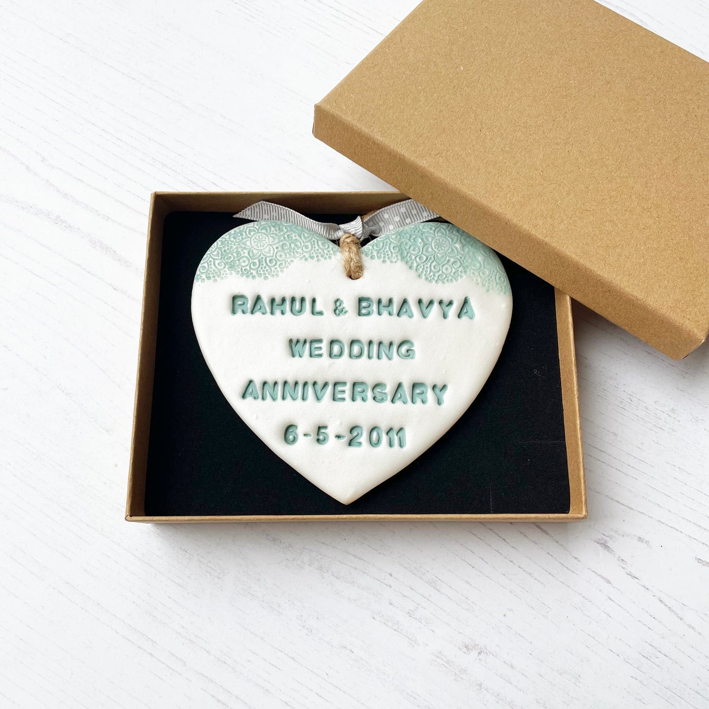 Personalised 13th wedding anniversary gift, pearlised white clay hanging heart with a sage green lace edge at the top of the heart, the heart is personalised with RAHUL & BHAVYA WEDDING ANNIVERSARY 6-5-2011 In a luxury Kraft brown gift box