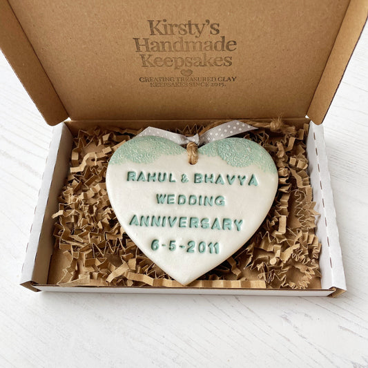 Personalised 13th wedding anniversary gift, pearlised white clay hanging heart with a sage green lace edge at the top of the heart, the heart is personalised with RAHUL & BHAVYA WEDDING ANNIVERSARY 6-5-2011 In a postal box with shredded Kraft brown zigzag paper