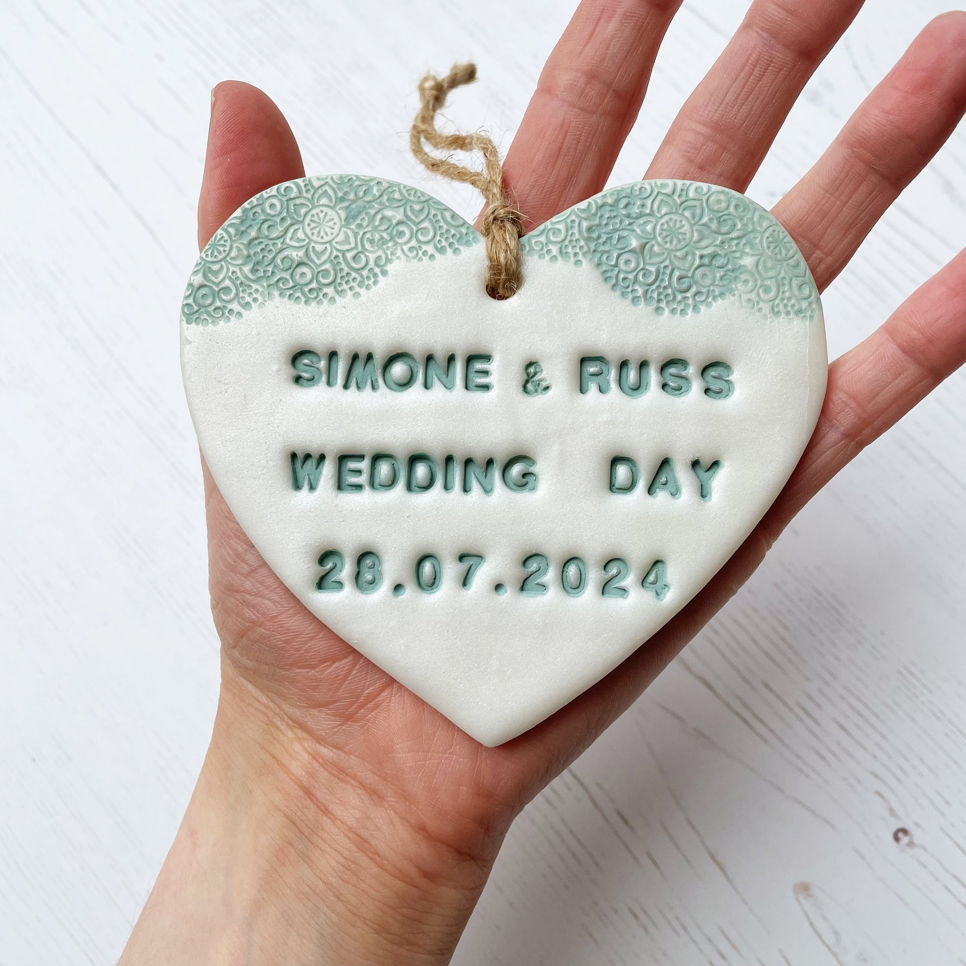 Personalised wedding gift, pearlised white clay heart with a sage green lace edge at the top of the heart with twine to hang, the heart is personalised with SIMONE & RUSS WEDDING DAY 28.07.2024