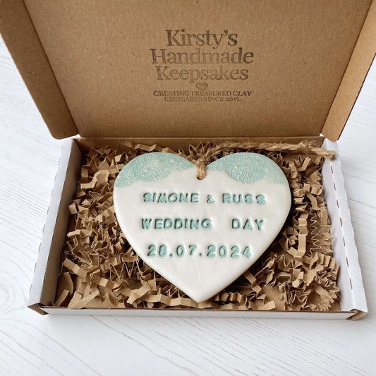 Personalised wedding gift, pearlised white clay heart with a sage green lace edge at the top of the heart with twine to hang, the heart is personalised with SIMONE & RUSS WEDDING DAY 28.07.2024