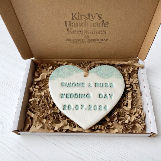 Personalised wedding gift, pearlised white clay heart with a sage green lace edge at the top of the heart with twine to hang, the heart is personalised with SIMONE & RUSS WEDDING DAY 28.07.2024 In a postal box with Kraft brown shredded zigzag paper 