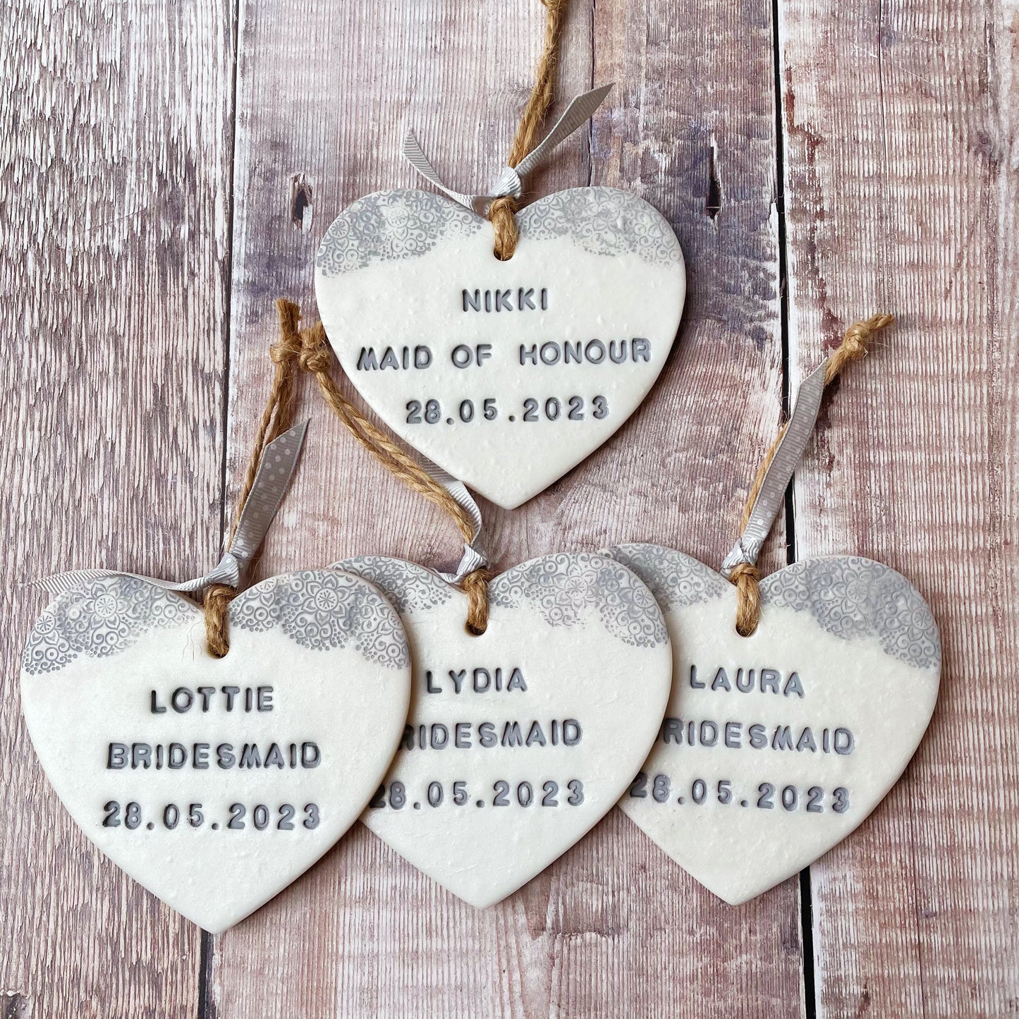 4 Personalised Bridesmaid thank you gifts, pearlised white clay hanging hearts with a grey lace edge at the top of the heart, the hearts are personalised with NIKKI MAID OF HONOUR 28.05.2023, LAURA BRIDESMAID 28.05.2023, LYDIA BRIDESMAID 28.05.2023 and LOTTIE BRIDESMAID 28.05.2023