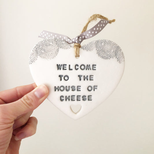 Personalised housewarming new home gift, pearlised white clay heart with a grey lace edge at the top of the heart and a heart cut out at the bottom with jute twine for hanging, the heart is personalised with WELCOME TO THE HOUSE OF CHEESE