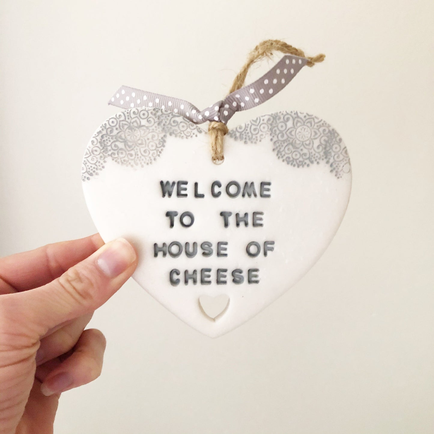Personalised housewarming new home gift, pearlised white clay heart with a grey lace edge at the top of the heart and a heart cut out at the bottom with jute twine for hanging, the heart is personalised with WELCOME TO THE HOUSE OF CHEESE