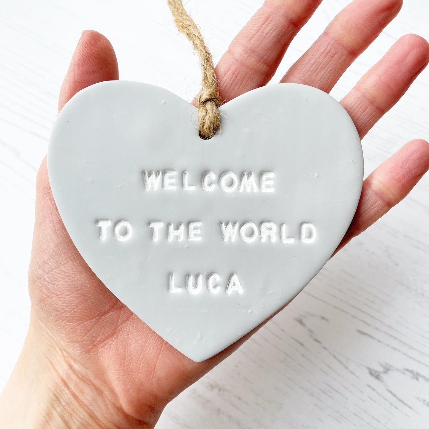 Personalised baby announcement heart sign, in grey clay hanging heart with a white writing, the heart is personalised with WELCOME TO THE WORLD LUCA