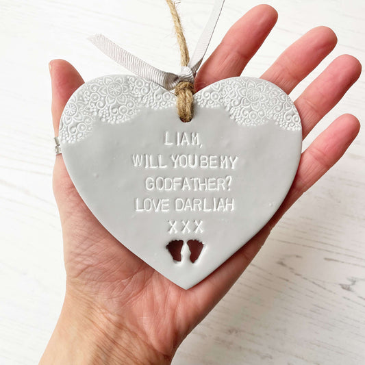 Personalised Godfather proposal gift, grey clay hanging heart with baby feet cut out of the bottom and white lace edge at the top of the heart, the heart is personalised with LIAM, WILL YOU BE MY GODFATHER? LOVE DARLIAH XXX