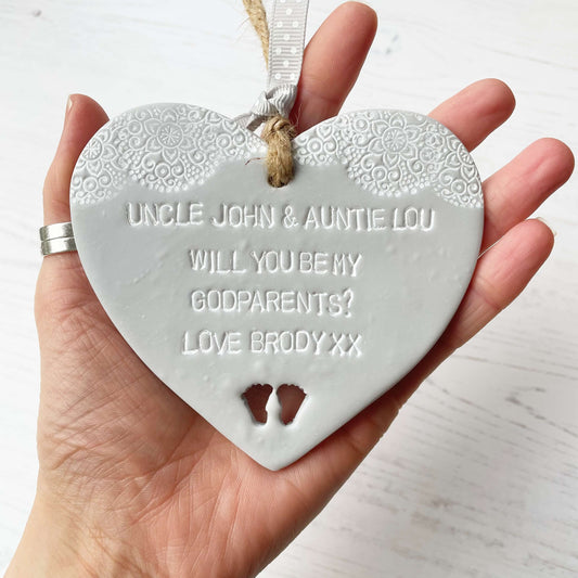 Personalised Godparents proposal gift, grey clay hanging heart with baby feet cut out of the bottom and white lace edge at the top of the heart, the heart is personalised with UNCLE JOHN & AUNTIE LOU WILL YOU BE MY GODPARENTS? LOVE BRODY XX