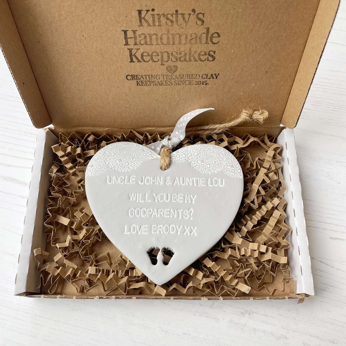 Personalised Godparents proposal gift, grey clay hanging heart with baby feet cut out of the bottom and white lace edge at the top of the heart, the heart is personalised with UNCLE JOHN & AUNTIE LOU WILL YOU BE MY GODPARENTS? LOVE BRODY XX
