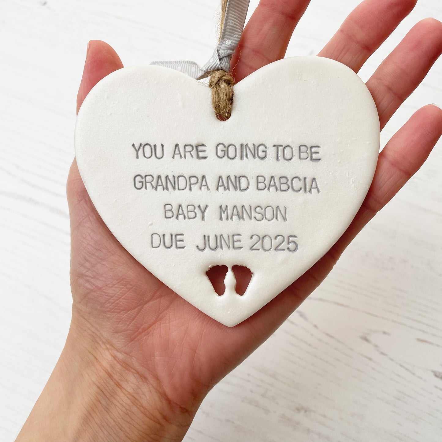 Pearlised white clay hanging heart with baby feet cut out of the bottom and grey personalisation, the heart is personalised with YOU ARE GOING TO BE GRANDPA AND BABCIA BABY MANSON DUE JUNE 2025