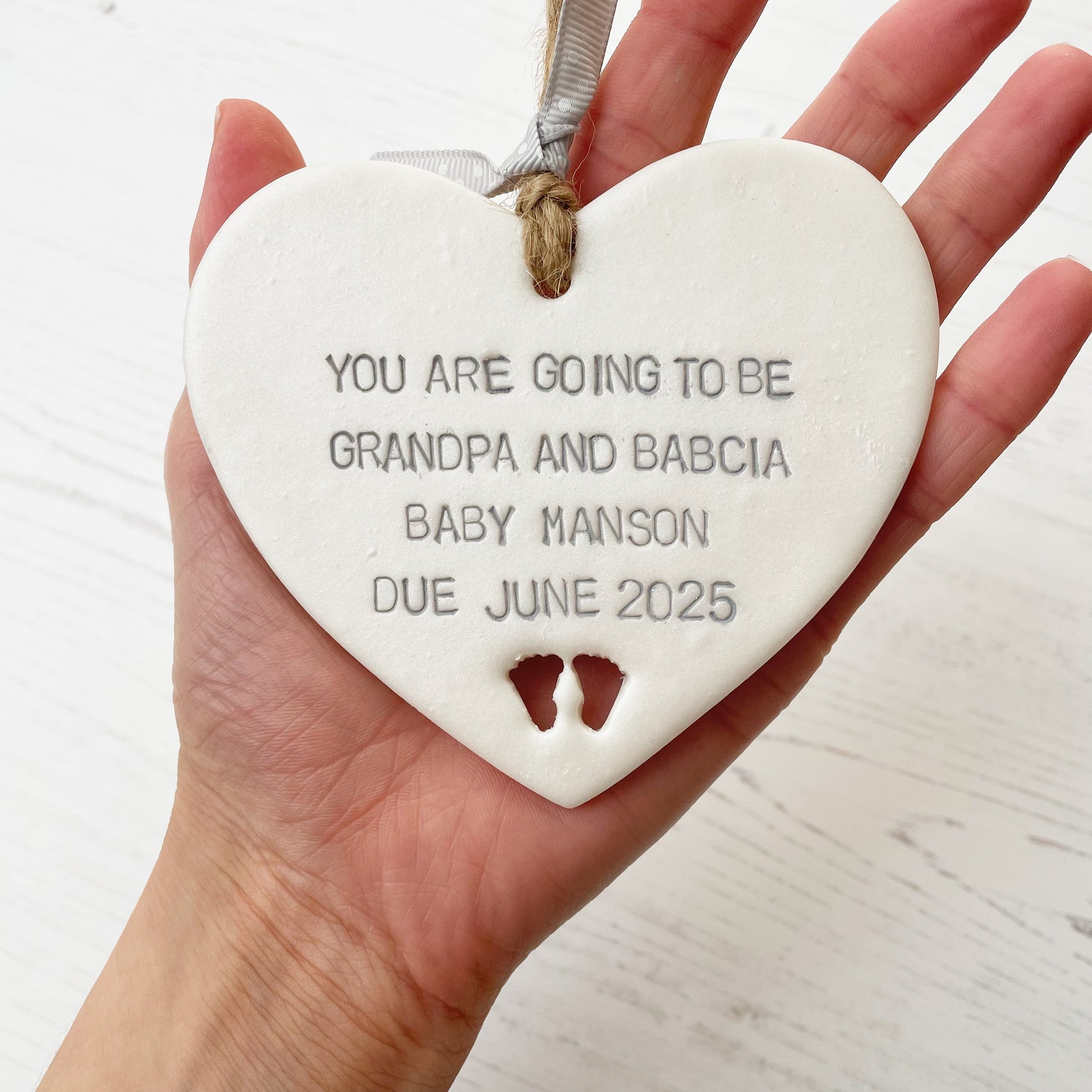 Pearlised white clay hanging heart with baby feet cut out of the bottom and grey personalisation, the heart is personalised with YOU ARE GOING TO BE GRANDPA AND BABCIA BABY MANSON DUE JUNE 2025