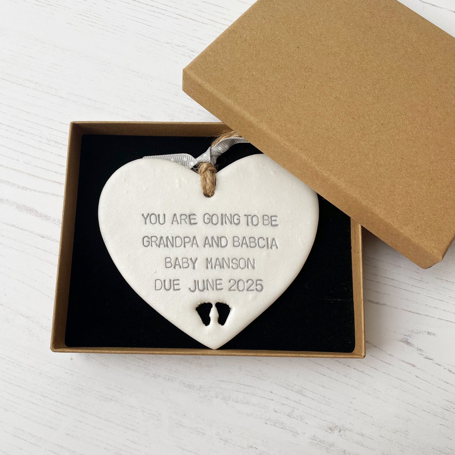 Pearlised white clay hanging heart with baby feet cut out of the bottom and grey personalisation, the heart is personalised with YOU ARE GOING TO BE GRANDPA AND BABCIA BABY MANSON DUE JUNE 2025