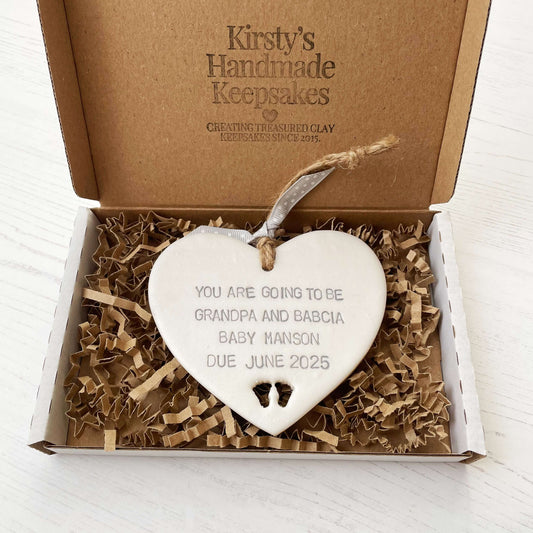 Pearlised white clay hanging heart with baby feet cut out of the bottom and grey personalisation, the heart is personalised with YOU ARE GOING TO BE GRANDPARENTS BABY MANSON DUE JUNE 2025