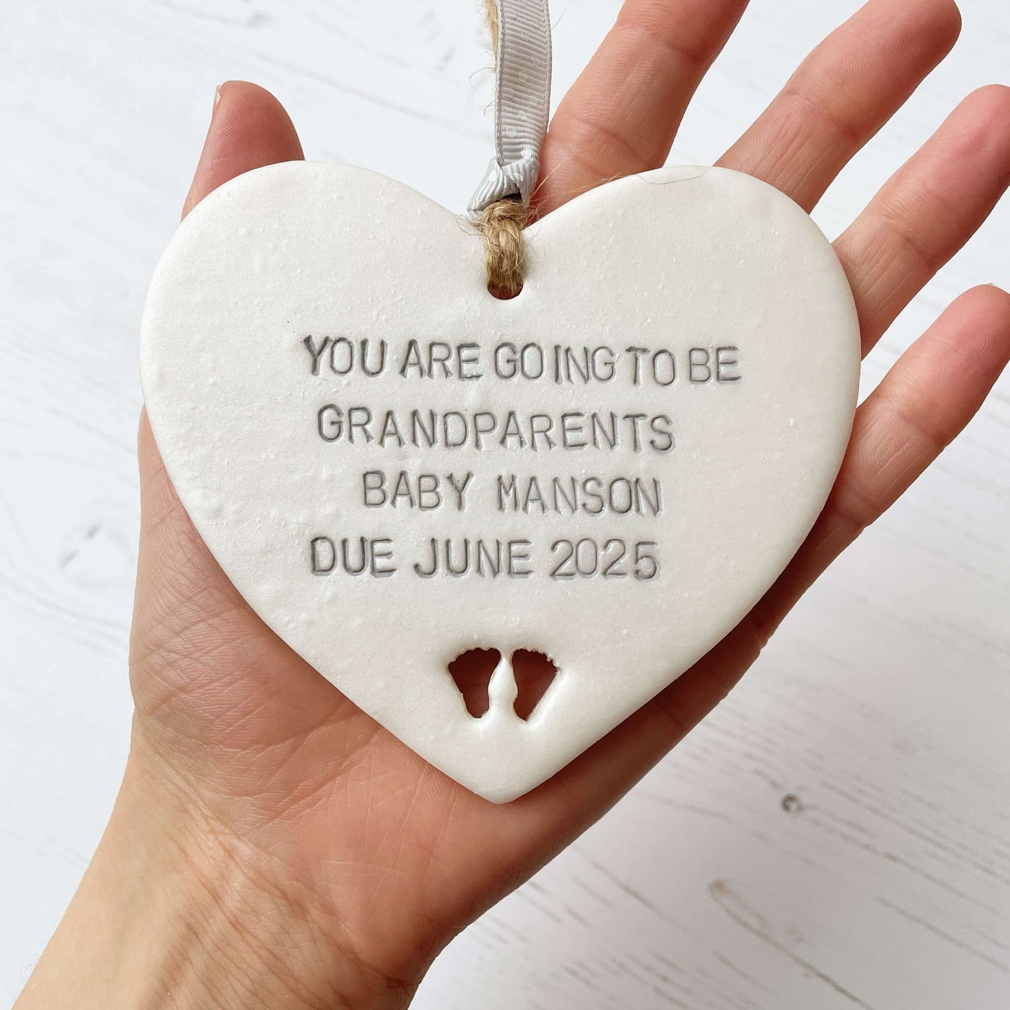 Pearlised white clay hanging heart with baby feet cut out of the bottom and grey personalisation, the heart is personalised with YOU ARE GOING TO BE GRANDPARENTS BABY MANSON DUE JUNE 2025