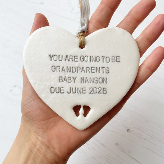 Pearlised white clay hanging heart with baby feet cut out of the bottom and grey personalisation, the heart is personalised with YOU ARE GOING TO BE GRANDPARENTS BABY MANSON DUE JUNE 2025