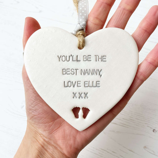 Pearlised white clay hanging heart pregnancy reveal with baby feet cut out of the bottom and grey personalisation, the heart is personalised with YOU’LL BE THE BEST NANNY, LOVE ELLE XXX