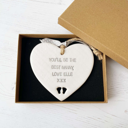 Pearlised white clay hanging heart pregnancy reveal with baby feet cut out of the bottom and grey personalisation, the heart is personalised with YOU’LL BE THE BEST NANNY, LOVE ELLE XXX