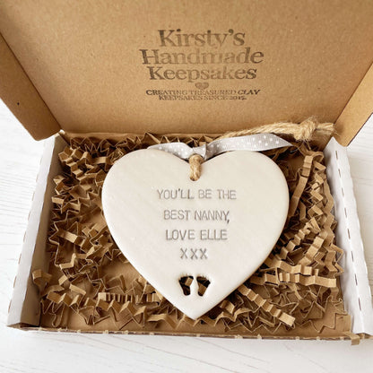 Pearlised white clay hanging heart pregnancy reveal with baby feet cut out of the bottom and grey personalisation, the heart is personalised with YOU’LL BE THE BEST NANNY, LOVE ELLE XXX
