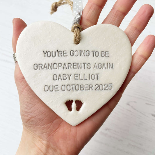 Pearlised white clay hanging heart with baby feet cut out of the bottom and grey personalisation, the heart is personalised with YOU’RE GOING TO BE GRANDPARENTS AGAIN BABY ELLIOT DUE OCTOBER 2025