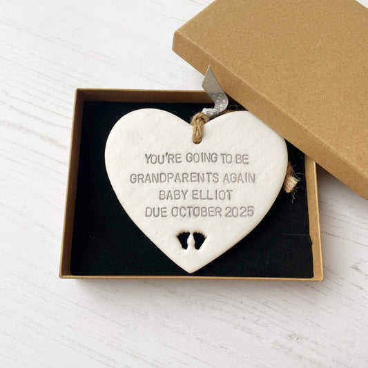 Pearlised white clay hanging heart with baby feet cut out of the bottom and grey personalisation, the heart is personalised with YOU’RE GOING TO BE GRANDPARENTS AGAIN BABY ELLIOT DUE OCTOBER 2025
