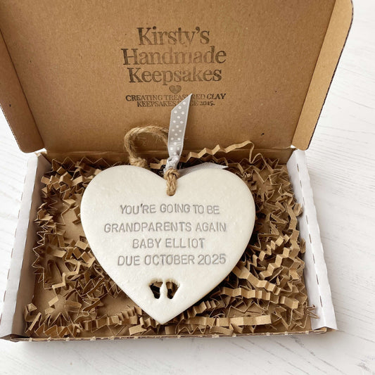 Pearlised white clay hanging heart with baby feet cut out of the bottom and grey personalisation, the heart is personalised with YOU’RE GOING TO BE GRANDPARENTS AGAIN BABY ELLIOT DUE OCTOBER 2025