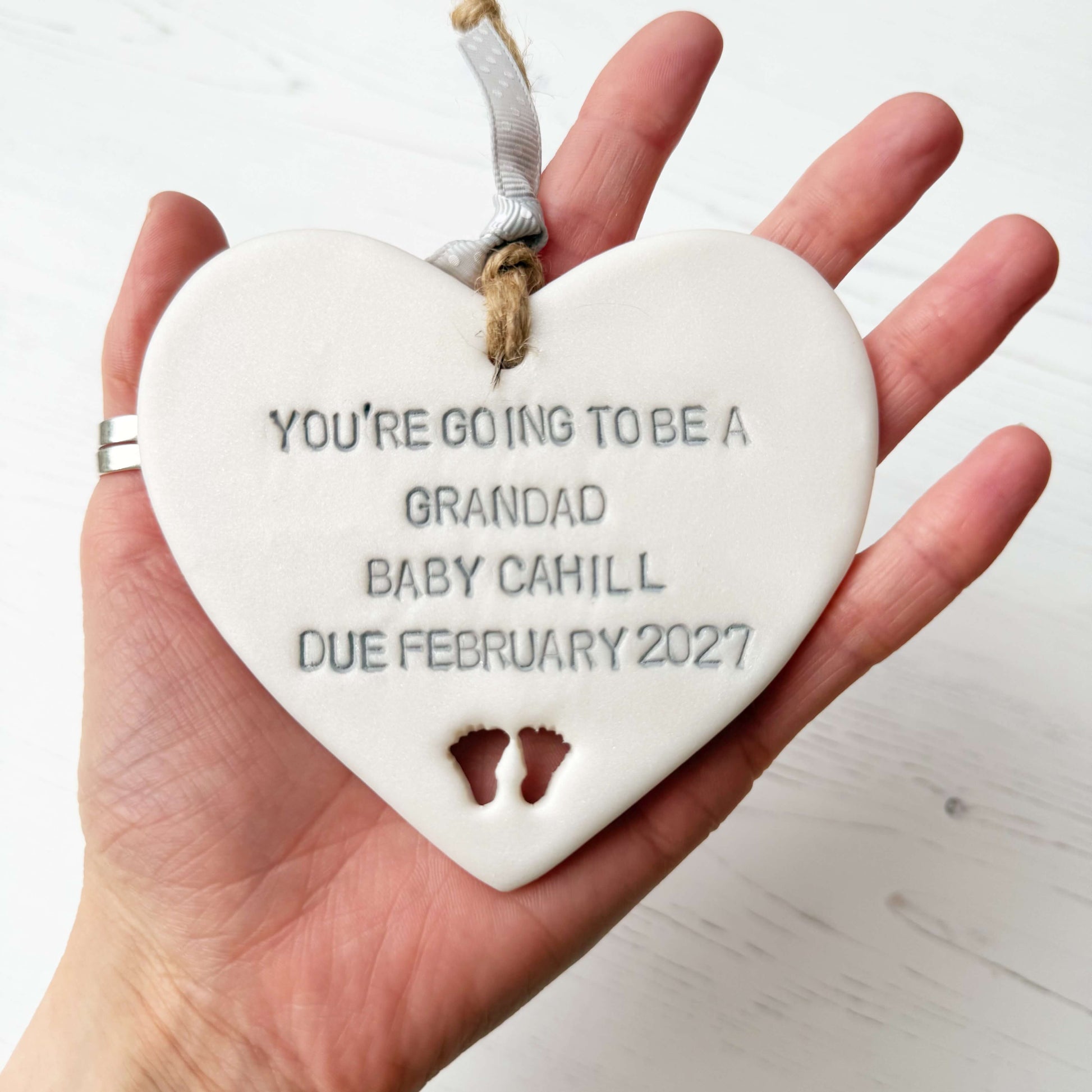 Pearlised white clay hanging heart with baby feet cut out of the bottom and grey personalisation, the heart is personalised with YOU’RE GOING TO BE A GRANDAD BABY CAHILL DUE FEBRUARY 2027