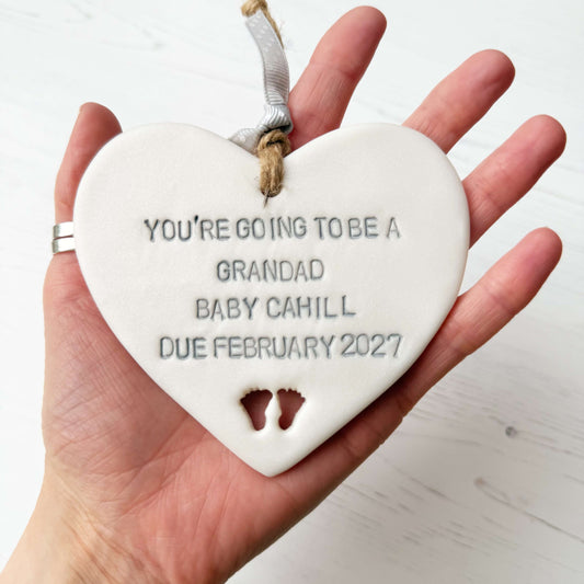 Pearlised white clay hanging heart with baby feet cut out of the bottom and grey personalisation, the heart is personalised with YOU’RE GOING TO BE A GRANDAD BABY CAHILL DUE FEBRUARY 2027