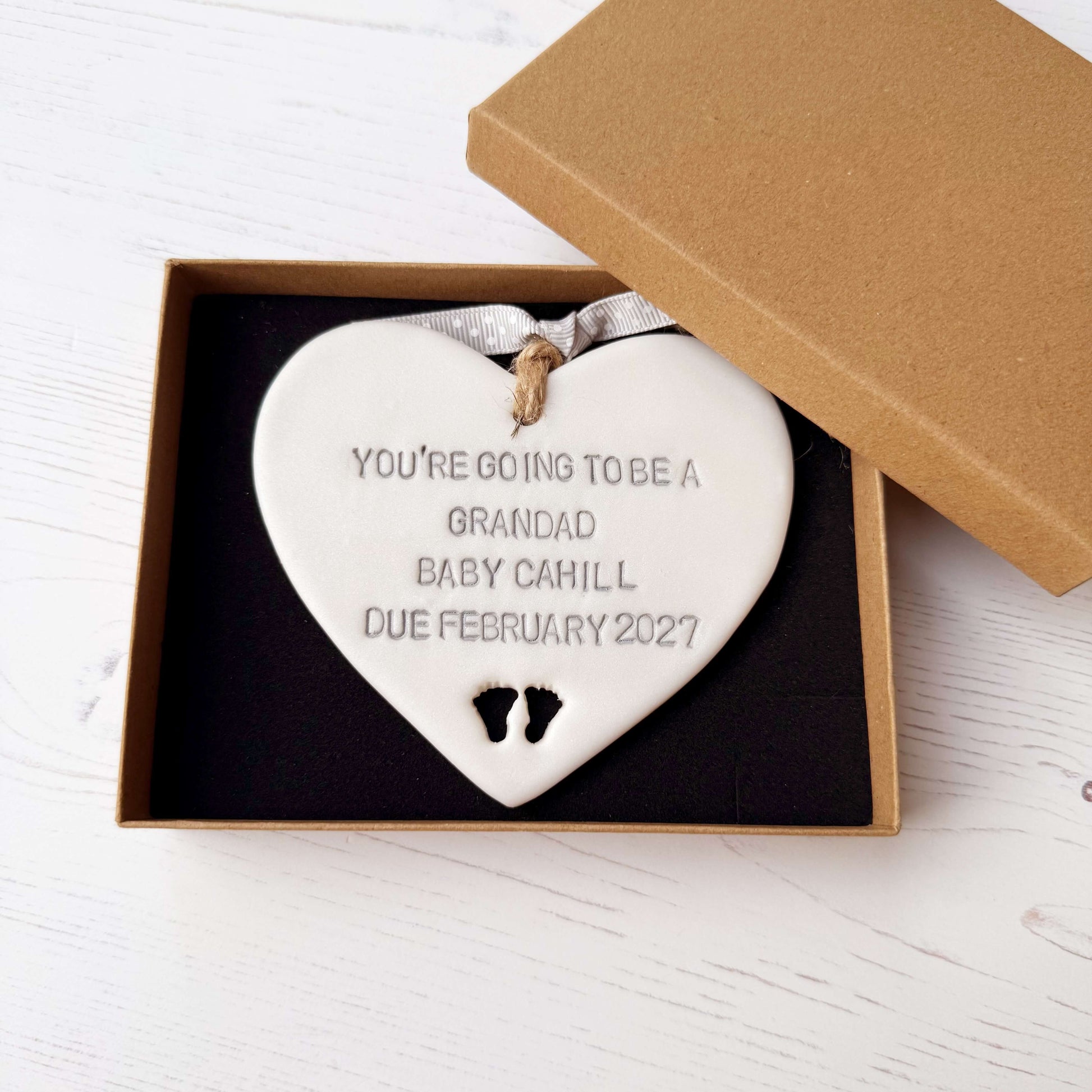 Pearlised white clay hanging heart with baby feet cut out of the bottom and grey personalisation, the heart is personalised with YOU’RE GOING TO BE A GRANDAD BABY CAHILL DUE FEBRUARY 2027