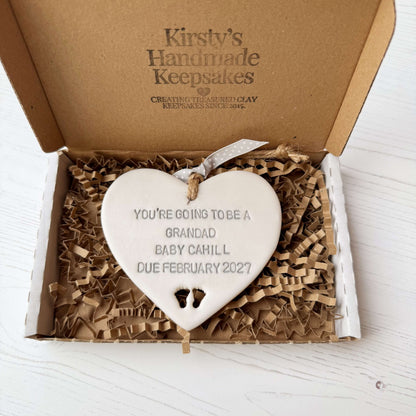 Pearlised white clay hanging heart with baby feet cut out of the bottom and grey personalisation, the heart is personalised with YOU’RE GOING TO BE A GRANDAD BABY CAHILL DUE FEBRUARY 2027