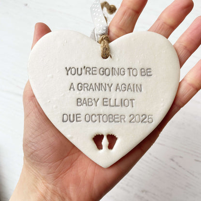 Pearlised white clay hanging heart with baby feet cut out of the bottom and grey personalisation, the heart is personalised with YOU’RE GOING TO BE A GRANNY AGAIN BABY ELLIOT DUE OCTOBER 2025