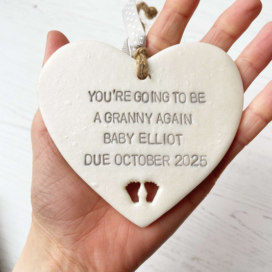 Pearlised white clay hanging heart with baby feet cut out of the bottom and grey personalisation, the heart is personalised with YOU’RE GOING TO BE A GRANNY AGAIN BABY ELLIOT DUE OCTOBER 2025
