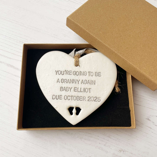 Pearlised white clay hanging heart with baby feet cut out of the bottom and grey personalisation, the heart is personalised with YOU’RE GOING TO BE A GRANNY AGAIN BABY ELLIOT DUE OCTOBER 2025