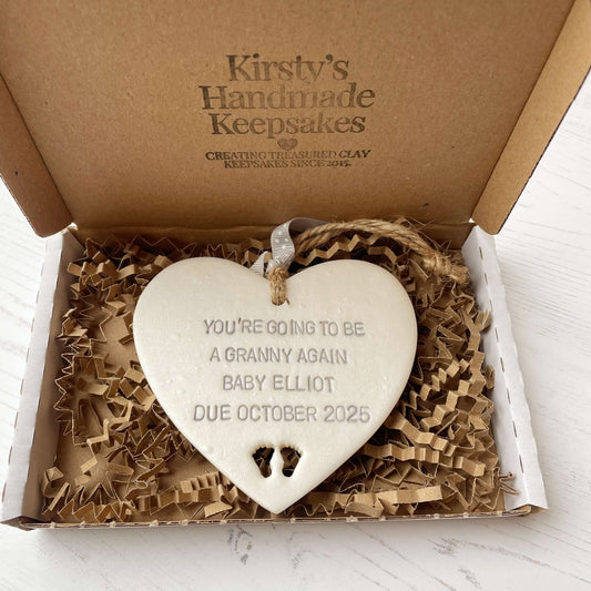 Pearlised white clay hanging heart with baby feet cut out of the bottom and grey personalisation, the heart is personalised with YOU’RE GOING TO BE A GRANNY AGAIN BABY ELLIOT DUE OCTOBER 2025
