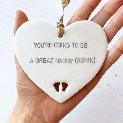 Pearlised white clay hanging heart with baby feet cut out of the bottom and grey personalisation, the heart is personalised with YOU’RE GOING TO BE GREAT NANNY (AGAIN)